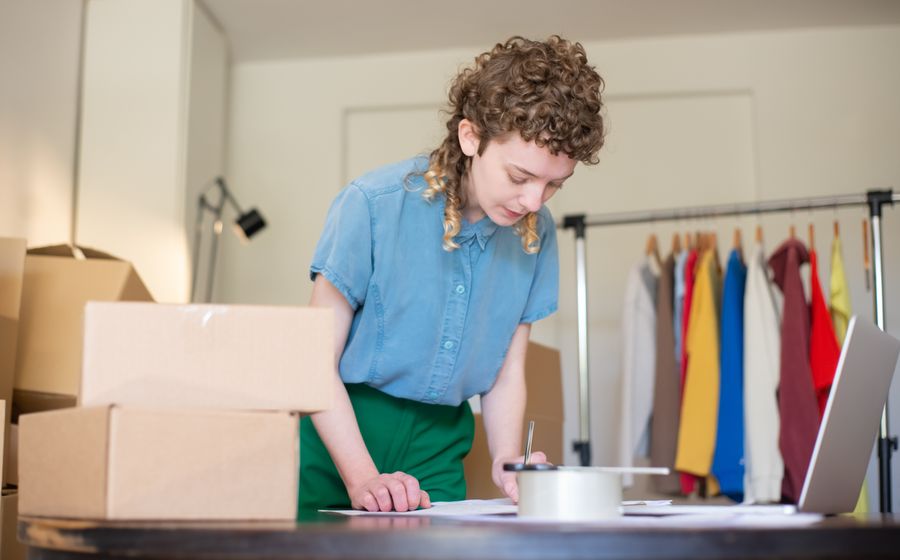Persona preparando pedidos en cajas junto a un portátil y un perchero de ropa.